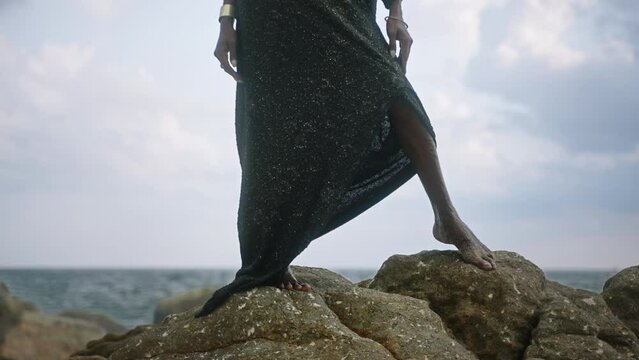 Godlike gender fluid person of color in luxury dress, jewelry stands on cliff peak at scenic ocean. Pride extravagant fashion model in epic outfit posing like goddess in tropics. Olymp concept.