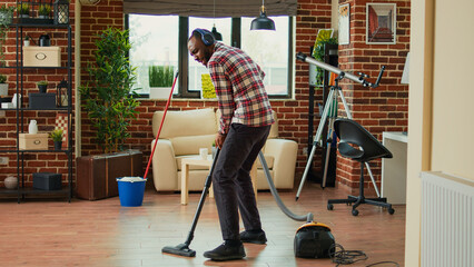 African american man dancing and vacuuming home floors, singing and listening to music on headset. Male person having fun spring cleaning, using vacuum to clean dust and dirt.