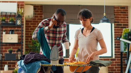 Young housewife ironing laundered clothes and feeling frustrated about doing chores alone, getting angry at husband. Stressed person throwing blouse at partner, being tired at home. Handheld shot.