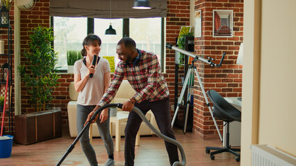 Funny cheerful people singing song and cleaning living room floors, using mop and vacuum cleaner at...