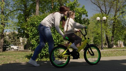 Fototapeta na wymiar happy family moments, playful girl, little daughter learns ride two-wheeled bicycle for first time, cardio, mom little daughter learn ride bike nature, child pedals wheels, happy family in the park