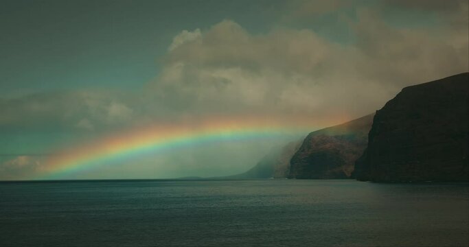 Dramatic stormy clouds on sky with vivid rainbow and dark ocean landscape. Igneous rocks Los Gigantes cliffs in Tenerife.