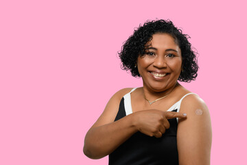 A woman smiles. She points to the vaccine sticker on her arm, on pink background
