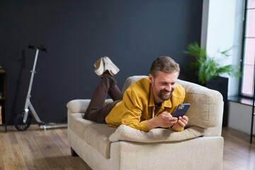 Smiling man enjoying time at home, sitting on sofa with phone. He appears handsome and content engages in texting or messaging on mobile device.