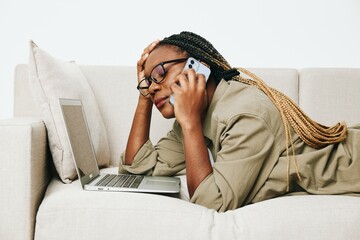 Happy african american woman business freelancer working sitting on sofa at home in laptop and phone, business calls and correspondence, light interior background.