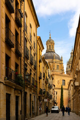 Scenic view of typical narrow cobbled street between medieval stone townhouses in Salamanca against backdrop of huge baroque dome of La Clerecia on spring day, Spain