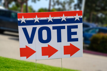 A VOTE sign at a polling place on green grass near a parking lot