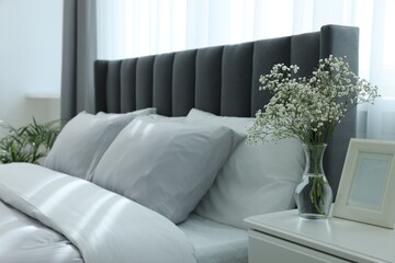 Vase with beautiful flowers on white bedside table near comfortable bed in room, space for text. Stylish interior