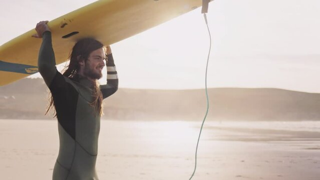 Young man carrying a surfboard walking on ocean beach. Surfer sports man getting ready to practice in the sea, catch a wave on a high tide. Active vacation evening or morning at the beach. 