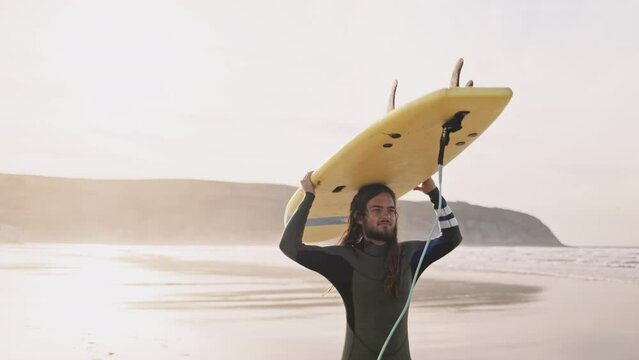 Young man carrying a surfboard walking on ocean beach. Surfer sports man getting ready to practice in the sea, catch a wave on a high tide. Active vacation evening or morning at the beach. 