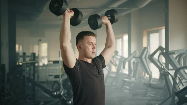 Medium shot of young athlete performing dumbbell shoulder press, standing in gym. Man holds dumbbells outside shoulders, then presses weight above head. High quality 4k footage