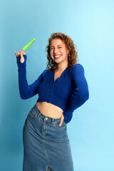 Portrait of young smiling girl in blue top and jeans skirt eating ice cream against blue studio background. Happiness. Concept of human emotions, youth culture, fashion, lifestyle