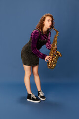 Portrait of emotional young girl in dress playing saxophone against blue studio background. Funny meme face. Concept of human emotions, youth culture, fashion, music lifestyle