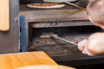 The chef cleaning the pizza oven. Raw pizza ready to bake. Cook in a blue apron in the kitchen. with a shovel in his hands. boxes for food delivery on background.