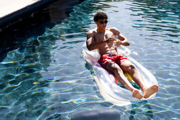 Biracial fit man in sunglasses relaxing on inflatable with cocktail in swimming pool, copy space