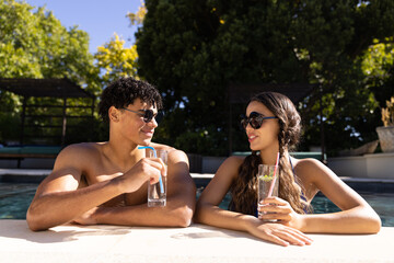 Happy diverse fit couple standing in swimming pool talking in the sun with cocktails