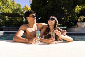 Happy diverse fit couple standing in swimming pool laughing in the sun with cocktails