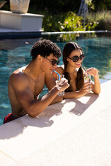 Happy diverse fit couple standing in swimming pool in the sun with cocktails, copy space
