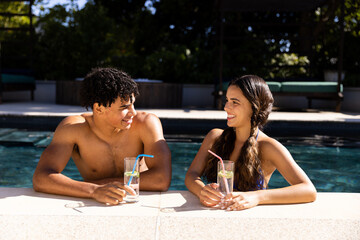 Happy diverse fit couple standing in swimming pool talking in the sun with cocktails