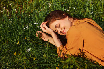 a calm woman with long red hair lies in a green field with yellow flowers, in an orange dress smiling pleasantly, closing her eyes from the bright summer sun
