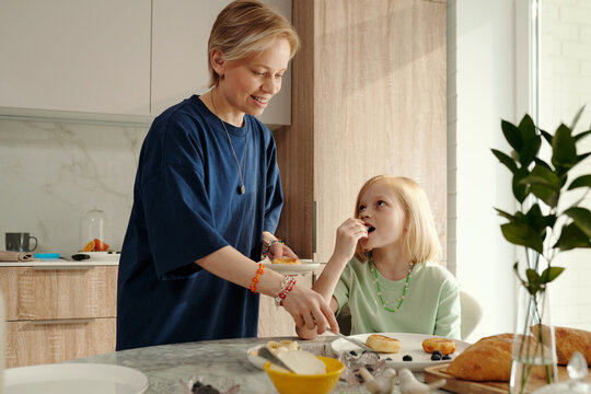 Modern mature Caucasian woman dishing out delicious quark pancakes for breakfast, her daughter sitting at table looking at her