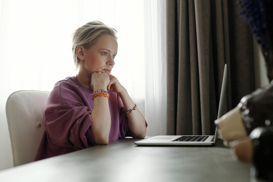 Modern Caucasian woman with short blond hair wearing casual outfit sitting at table at home watching video on laptop