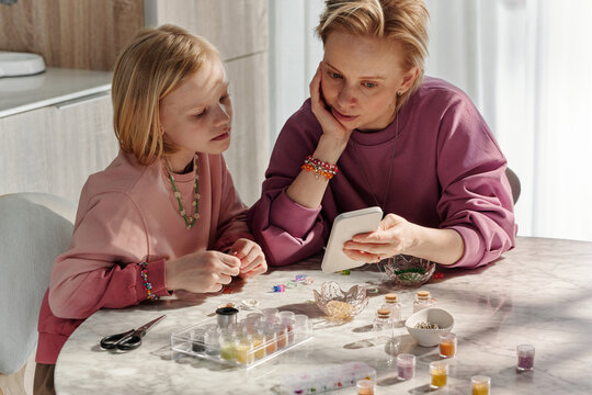 Modern Caucasian woman and her preteen daughter spending time together at home choosing design 
of bead accessories scrolling pictures in Internet