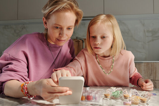 Creative Caucasian woman and her daughter spending weekend together at home choosing design of bead accessories scrolling pictures in Internet