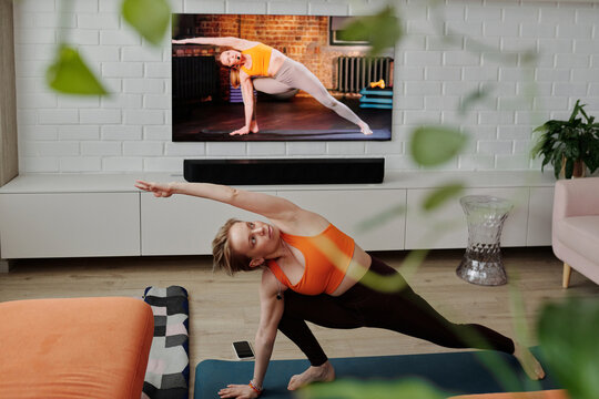 Modern Caucasian woman having yoga workout at home doing stretching and balancing asana with fitness trainer on TV screen