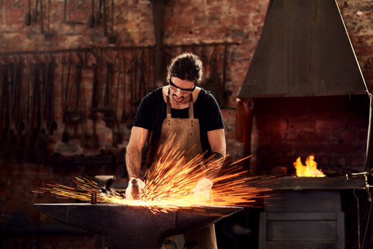 Blacksmith, man and metal workshop and sparks, manual forge working on hot steel with hammer, and fire. Worker, person welding or iron tools manufacturing and expert, trade and dark workspace