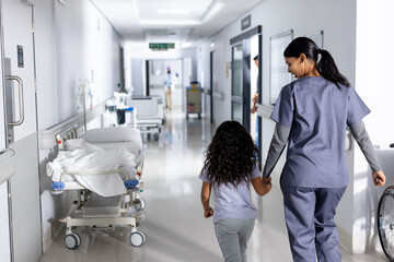 Smiling biracial female doctor and girl patient holding hands walking in hospital corridor