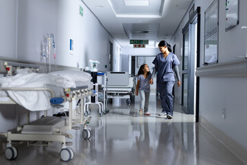 Biracial female doctor and girl patient holding hands, walking and talking in hospital corridor