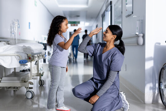 Happy kneeling biracial female doctor and girl patient high fiving in corridor at hospital