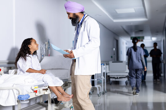 Happy sikh male doctor in turban and biracial girl patient talking in hospital corridor, copy space