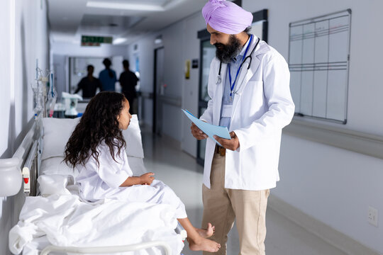Happy sikh male doctor in turban and biracial girl patient talking in corridor at hospital