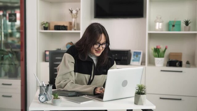 The young charismatic woman, a brunette, sits in an office at a desk in front of a laptop and works diligently and devotedly. She is engrossed in her work and mumbles something to herself.
