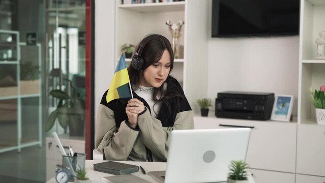 The young charismatic woman, a brunette, sits in an office in front of a laptop and communicates through video conferencing.She holds a Ukrainian flag.The woman is wearing wireless headphones.