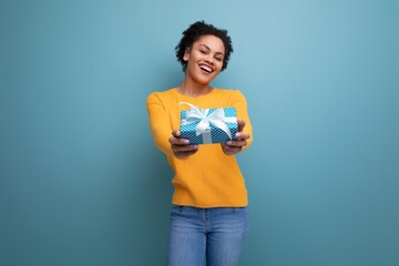 happy young hispanic woman with black curly hair in a yellow sweater holds out a gift box