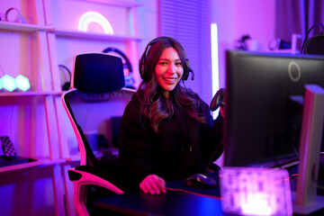 Asian pretty streamer girl smile and hold controller for game battle play while sitting comfort ergonomic chair with neon sign background in entertainment gamer room