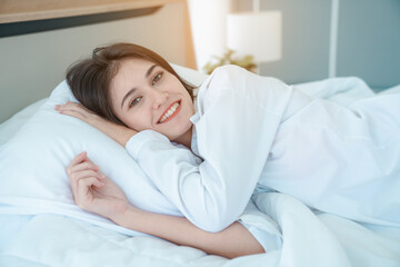 Fototapeta na wymiar Photo of young happy woman in pajama stretching her arms and smiling while sitting on bed after sleep