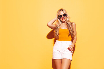 Young beautiful smiling blond female in trendy summer shorts clothes. Sexy carefree woman posing near yellow wall in studio. Positive model having fun. Cheerful and happy. Going crazy. In sunglasses