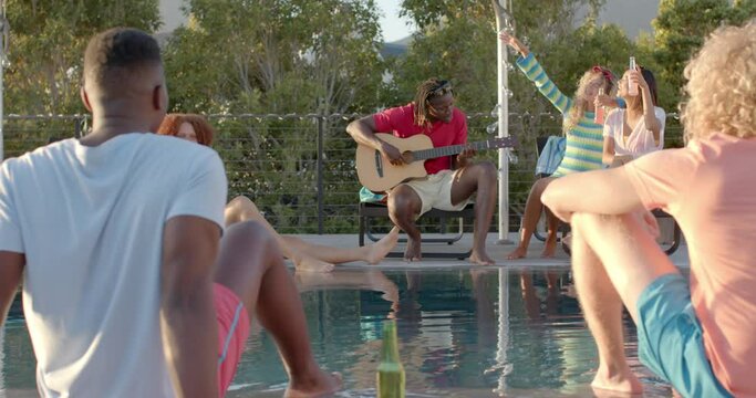 Happy diverse friends with drinks playing guitar at pool party in slow motion