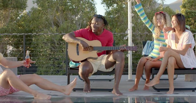 Happy diverse friends with drinks playing guitar at pool party in slow motion