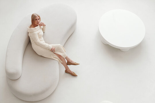 Horizontal from above view of elegant senior woman wearing fashionable dress sitting on white sofa in modern living room