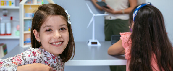 Portrait of smiling schoolgirl looking at camera in ecology classroom while female teacher explaining lesson to students on background. Renewable energies education concept.