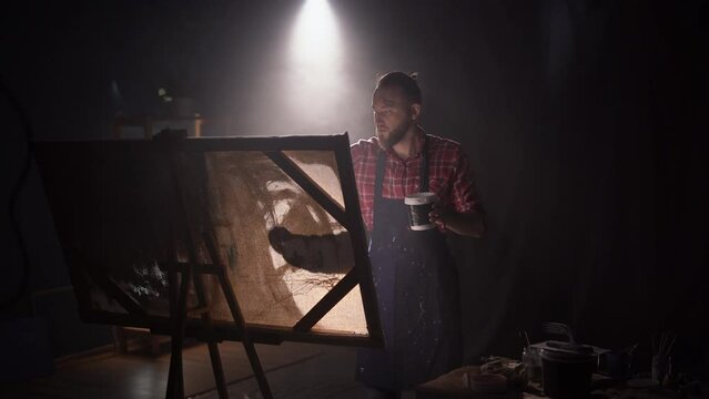 Male artist using paint brush creates of the acrylic painting. Creative studio with large canvas. Side view