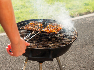 Cooking marinated chicken in a metal basket on small round grill. Cook hand out of focus. Sumer...