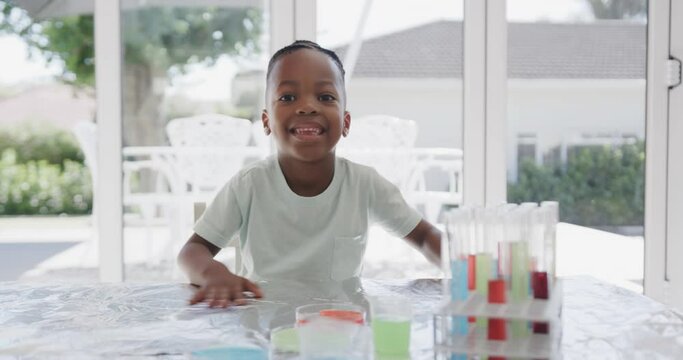 Portrait of african american boy sitting at table with test tubes and smiling, in slow motion