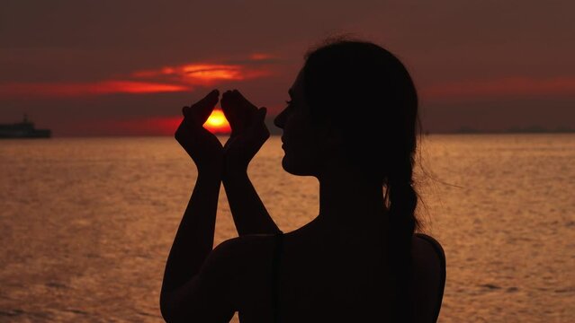Back view silhouette woman at sunset, close up making figure with hands holding inside the sun, symbol of peace and meditation. Happy woman looking at sea sunset, hands covers the sun before sunset