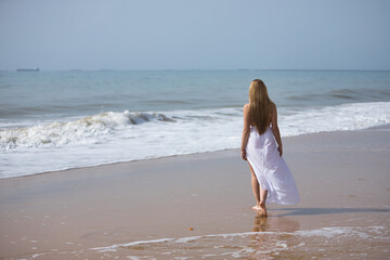 Fototapeta na wymiar Young, beautiful, blonde woman in white dress, walking on a lonely beach, relaxed and calm, seen from the back. Concept peace, tranquility, solitude, finding oneself.